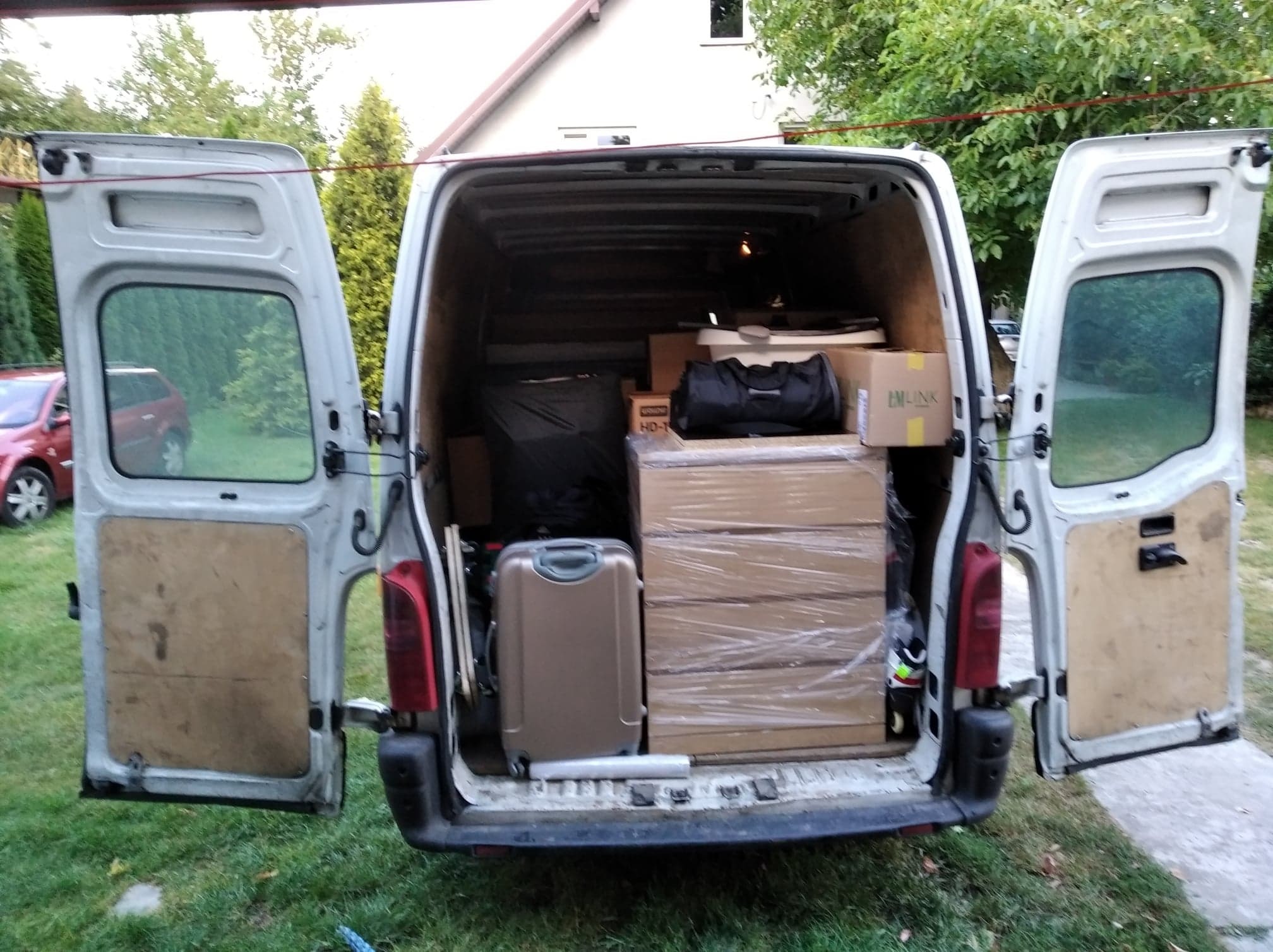Wnętrze białego busa z otwartymi drzwiami załadowane kartonami owiniętymi folią, walizką i torbami, widok z tyłu na trawnik i dom.