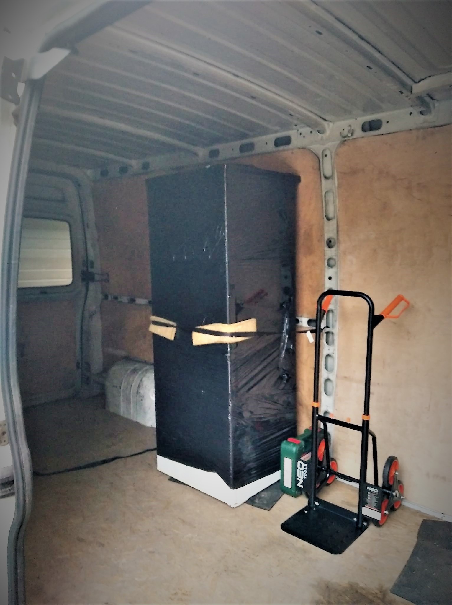 Wnętrze furgonetki z zabezpieczonym meblem owiniętym folią, obok wózek transportowy z trzema kołami po każdej stronie i kanister z olejem. Ściany furgonetki wyłożone płytami ze sklejki.