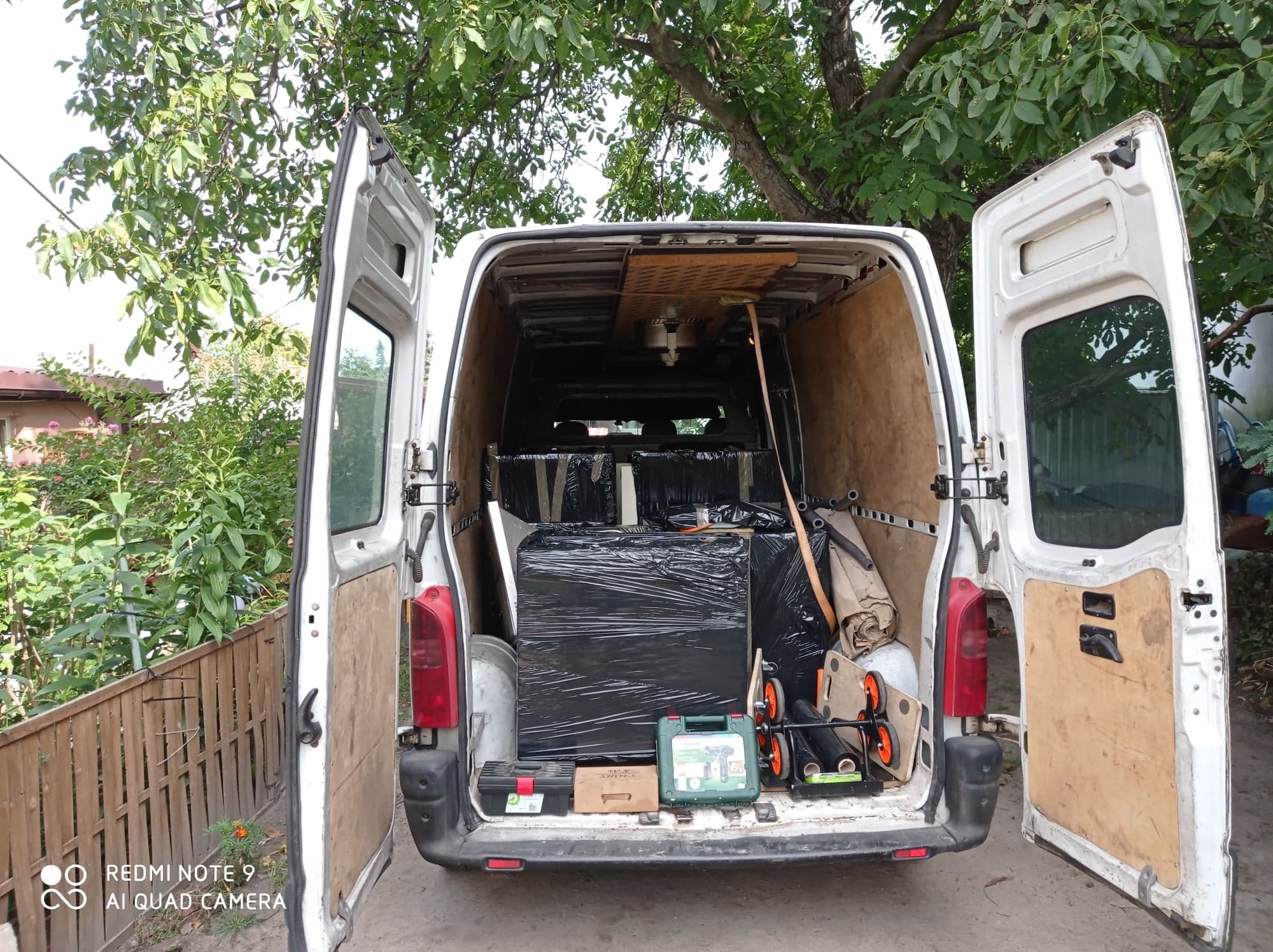 Otwarty biały bus załadowany meblami owiniętymi w czarną folię stretch, narzędzia w skrzynkach i akcesoria do transportu, widok z tyłu na tle zieleni.