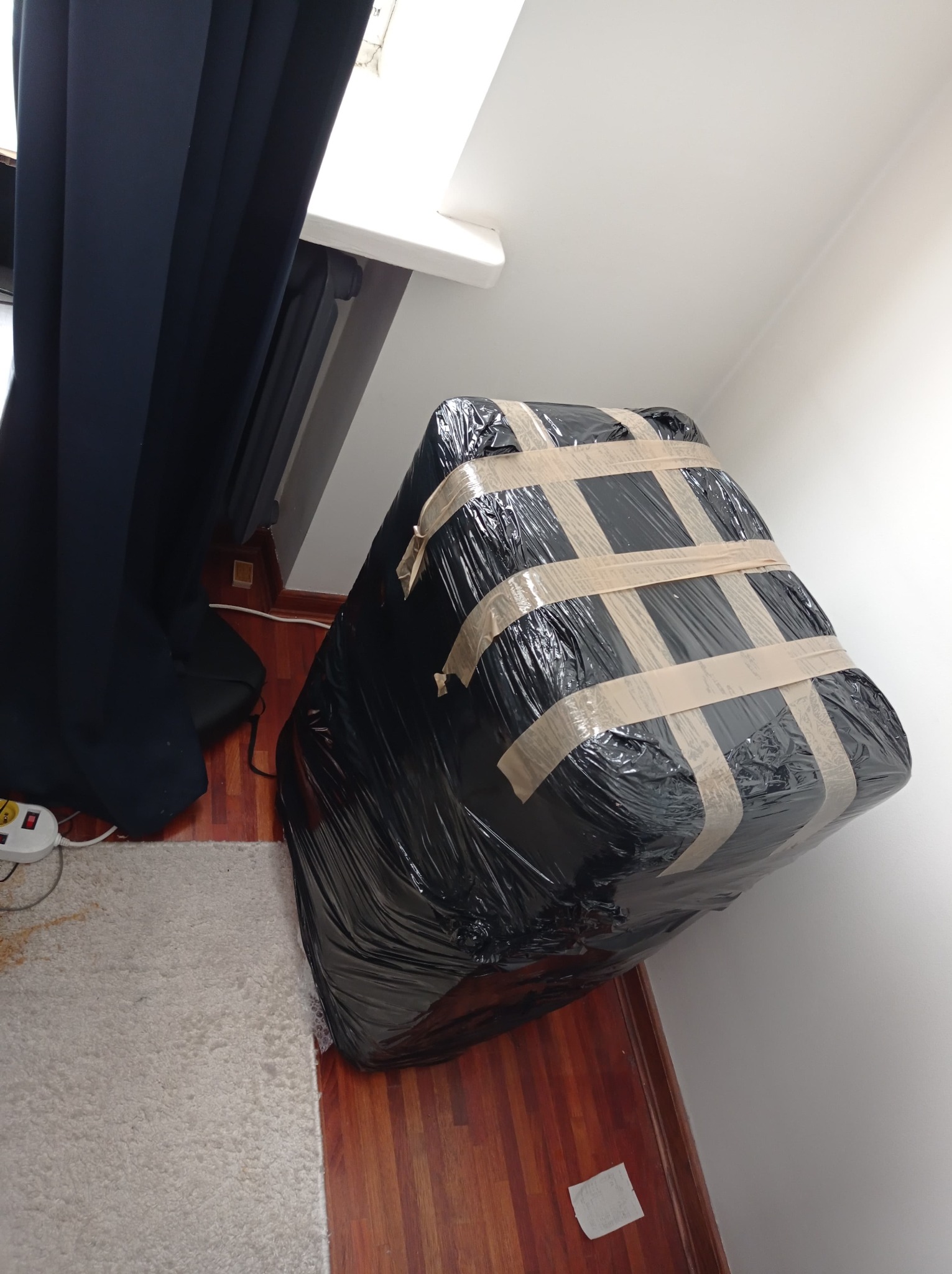 Spakowany i oklejony taśmą mebel w czarnej folii przygotowany do transportu w pomieszczeniu z zasłoniętym oknem.