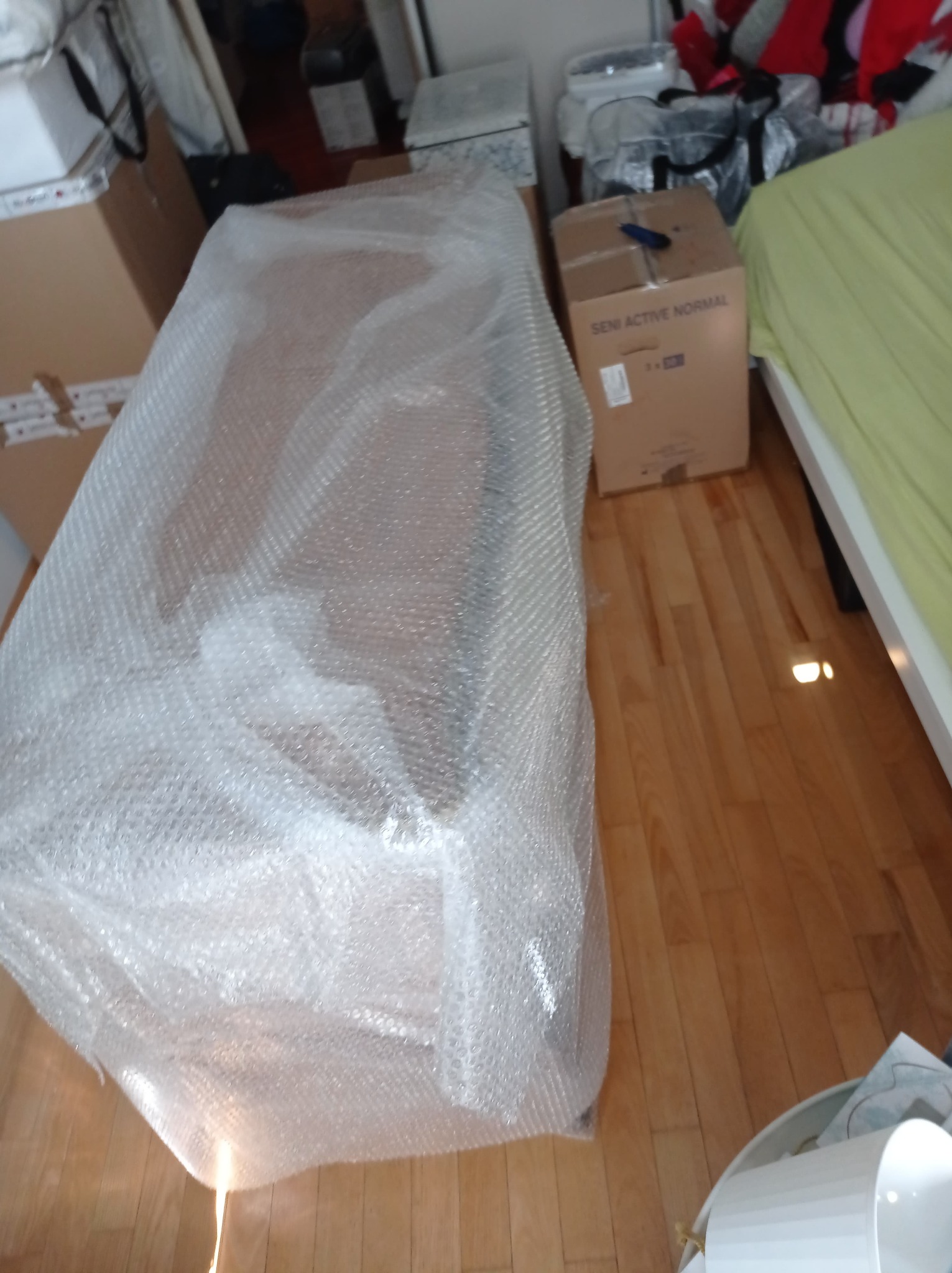 Meble zabezpieczone folią bąbelkową podczas transportu, karton z napisem 'SENI ACTIVE NORMAL' obok łóżka z zieloną pościelą, drewniana podłoga.