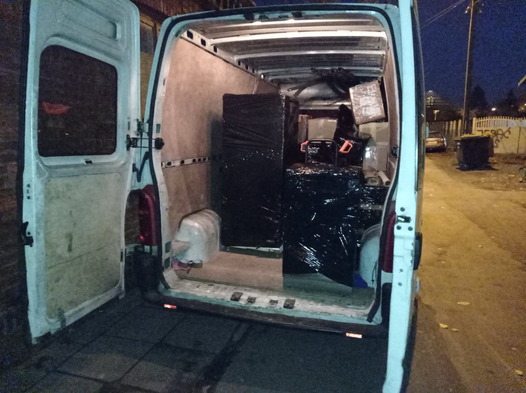 Wnętrze białego busa załadowane meblami i kartonami zabezpieczonymi czarną folią stretch, widoczne otwarte tylne drzwi i fragment ulicy w tle.