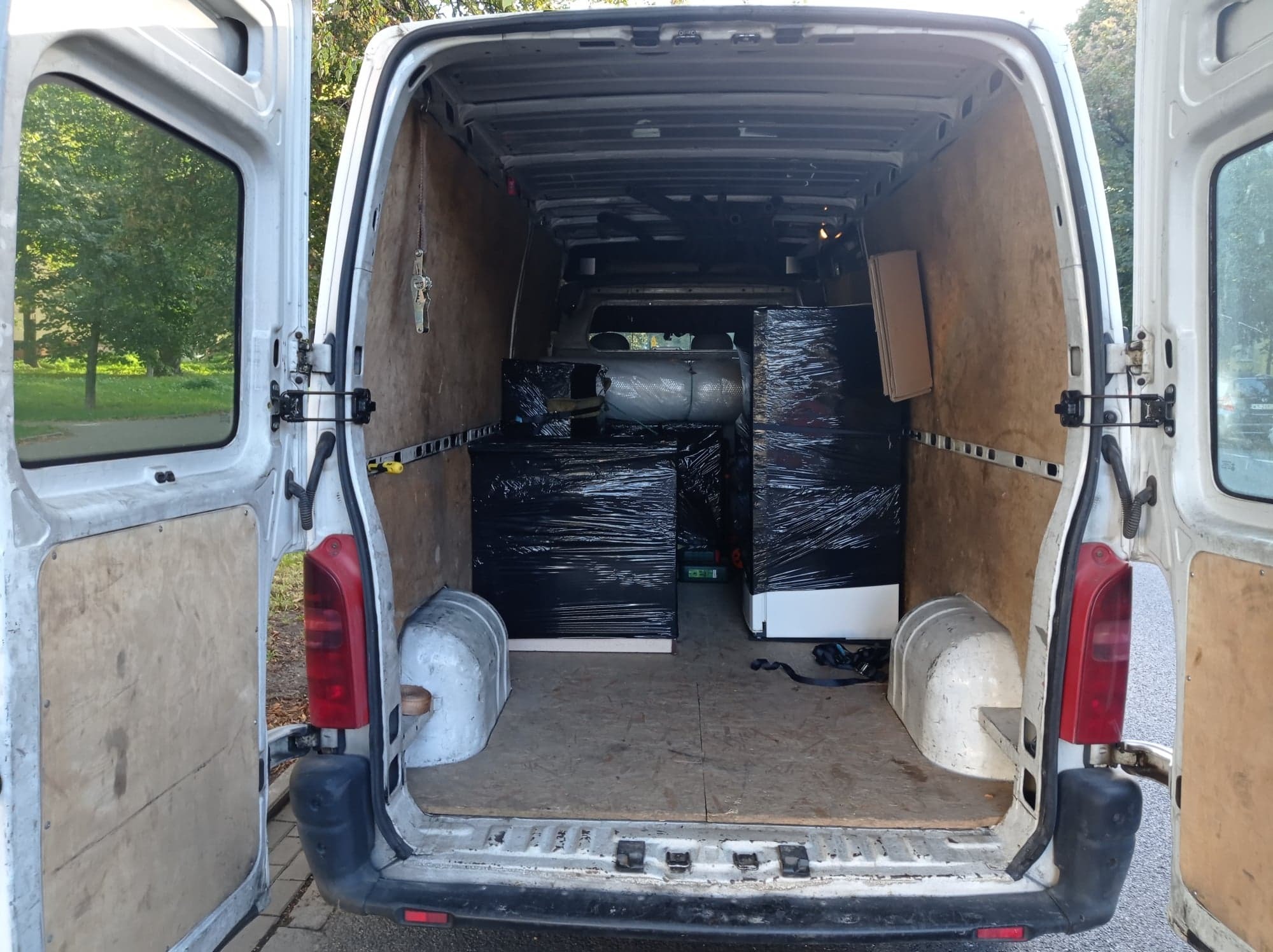 Wnętrze białego busa z otwartymi drzwiami, załadowane meblami owiniętymi czarną folią stretch, widok z tyłu pojazdu.