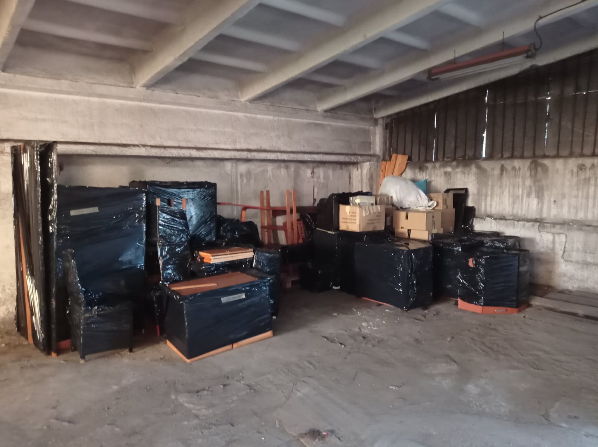 Spakowane meble i kartony w czarnej folii stretch, przygotowane do transportu, składowane w magazynie o betonowych ścianach i suficie.