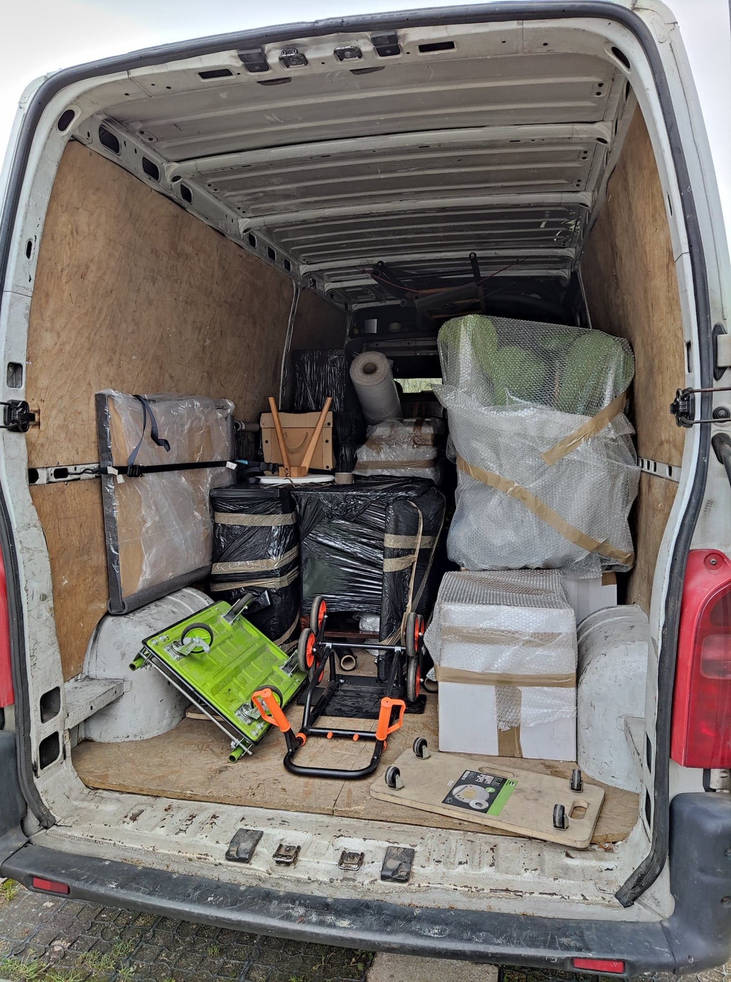 Wnętrze białego furgonetki wypełnione po brzegi zapakowanymi przedmiotami, w tym meblami owiniętymi folią bąbelkową, kartonami i wózkiem transportowym w kolorze limonkowym.