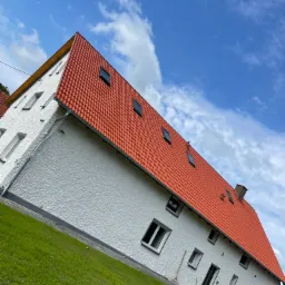 Dom z nowym, czerwonym dachem z dachówki ceramicznej i oknami dachowymi, widok z poziomu gruntu na tle błękitnego nieba z chmurami.