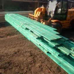 Stos impregnowanych na zielono desek na ziemi obok żółtej ładowarki teleskopowej i drewnianej konstrukcji w tle.