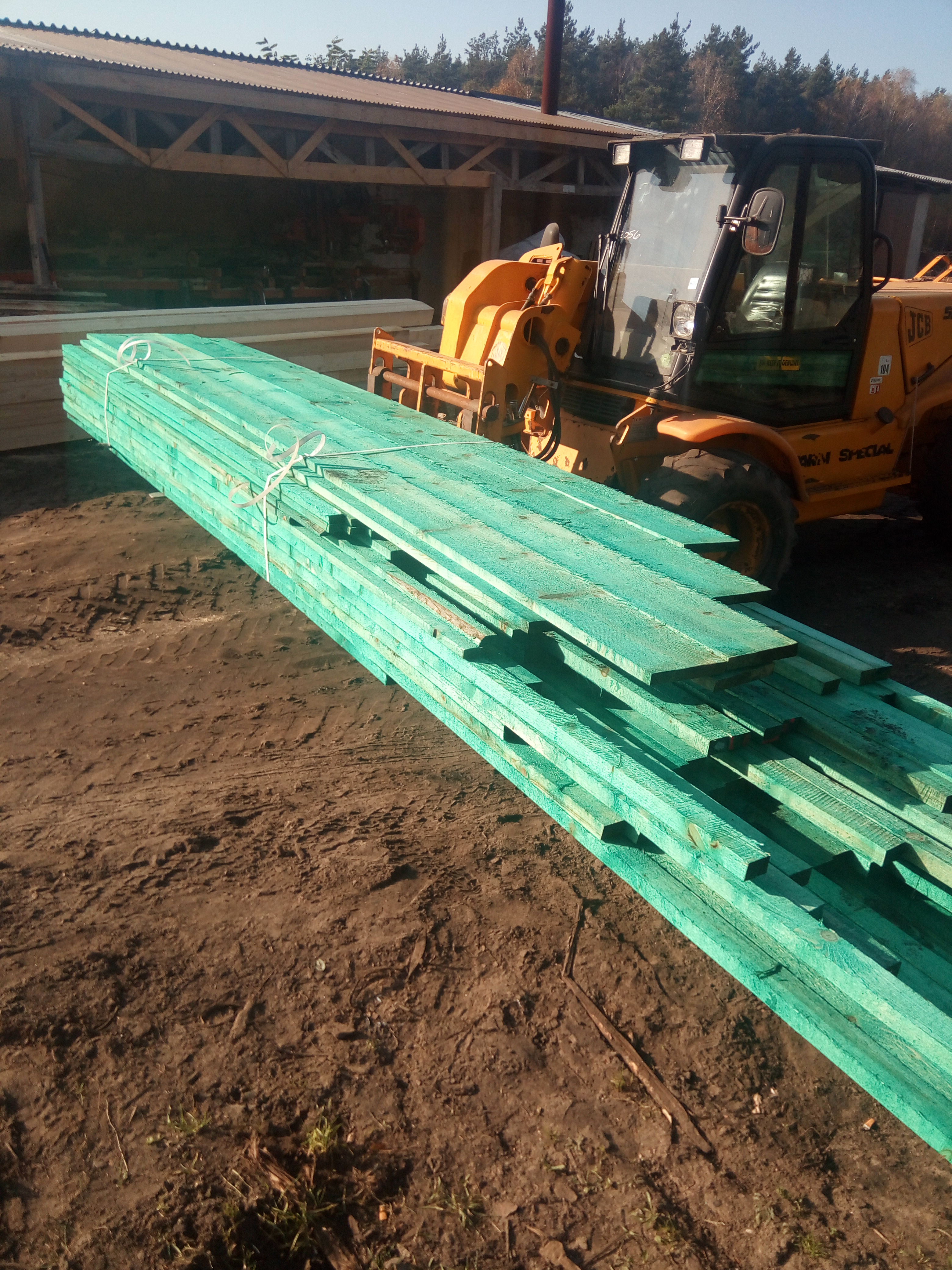 Stos impregnowanych na zielono desek na ziemi obok żółtej ładowarki teleskopowej i drewnianej konstrukcji w tle.