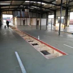 Wnętrze hali z betonową posadzką w trakcie wykonywania, widoczny pracownik szlifujący powierzchnię, odkryty kanał technologiczny z drewnianym szalunkiem i kratka odpływowa.