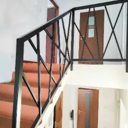 Klatka schodowa z nową, czarną, metalową balustradą o geometrycznym wzorze, widok z dołu na górę, z widocznymi drzwiami na różnych poziomach.