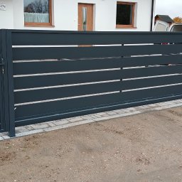 Adam Kleba - Nowoczesna, przesuwna brama posesyjna w kolorze grafitowym z poziomymi szczeblami, widoczna z zewnątrz. Obok furtka i fragment ogrodzenia z siatki.