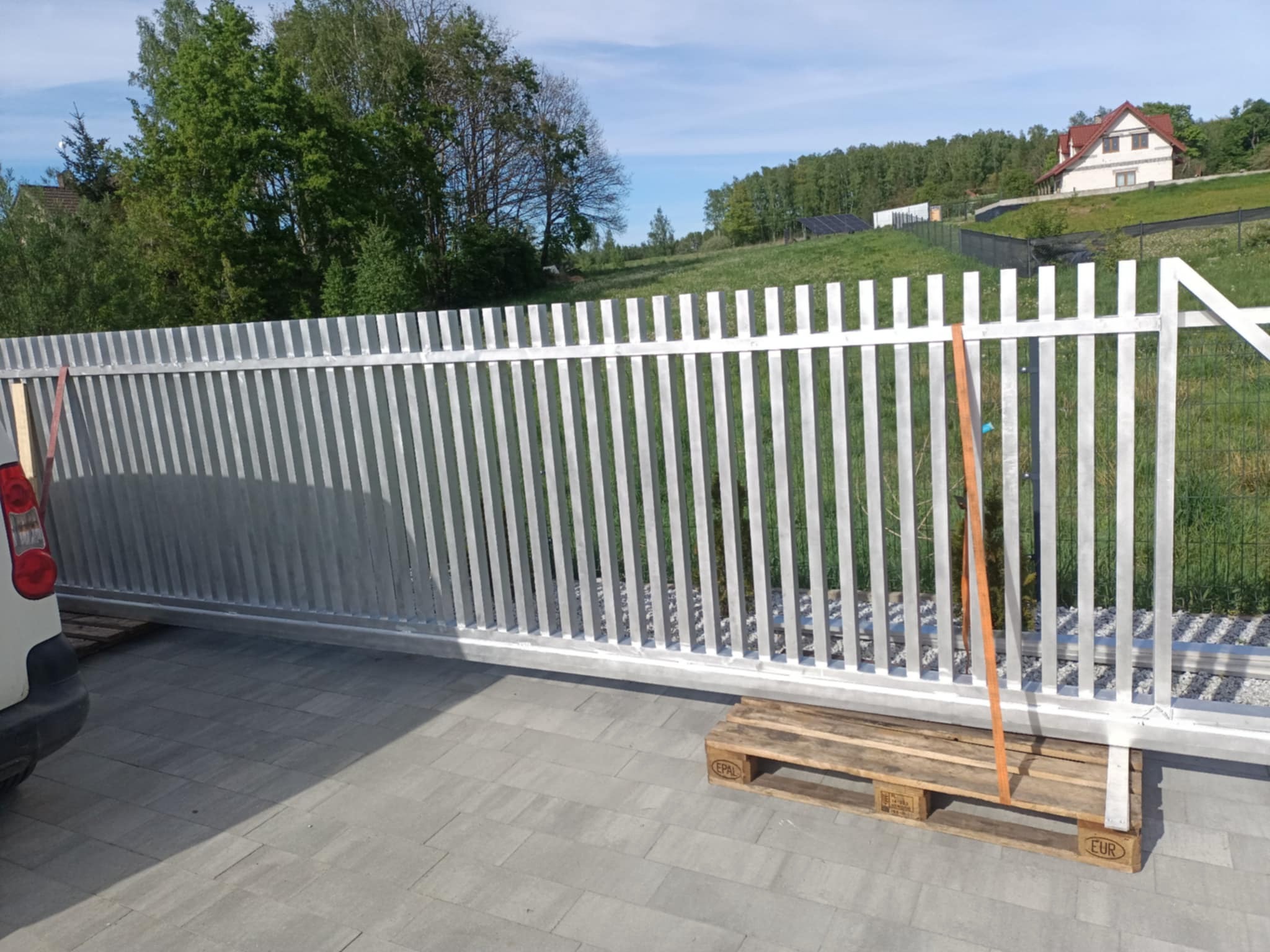 Nowa, srebrna, przesuwna brama metalowa z pionowymi szczeblami, umieszczona na palecie EUR, z widocznym fragmentem samochodu po lewej stronie, na tle zieleni i zabudowań.