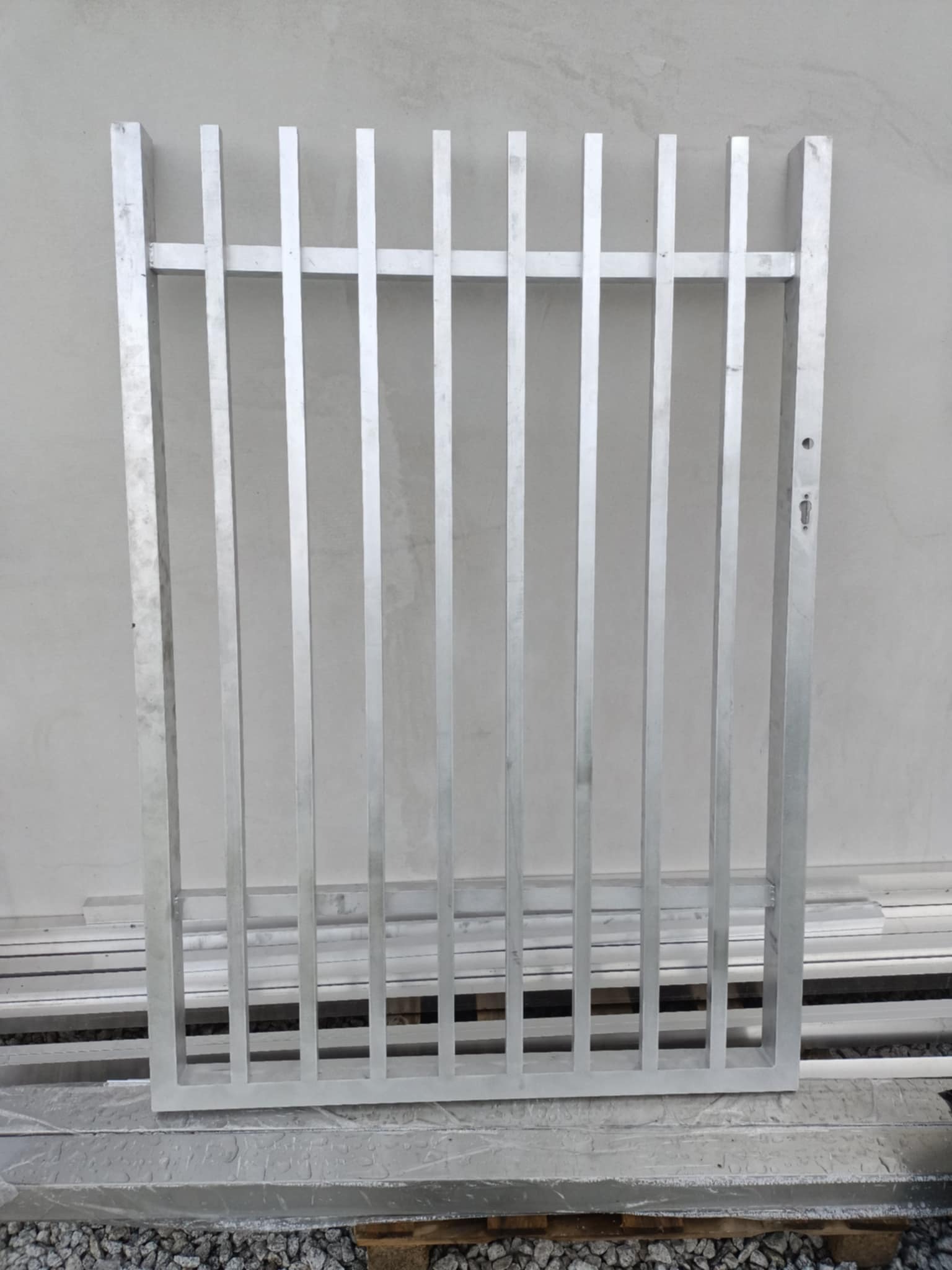Surowa, aluminiowa rama ogrodzeniowa z pionowymi szczeblami, widoczne ślady obróbki i spawania, leżąca na palecie na tle betonowej ściany.