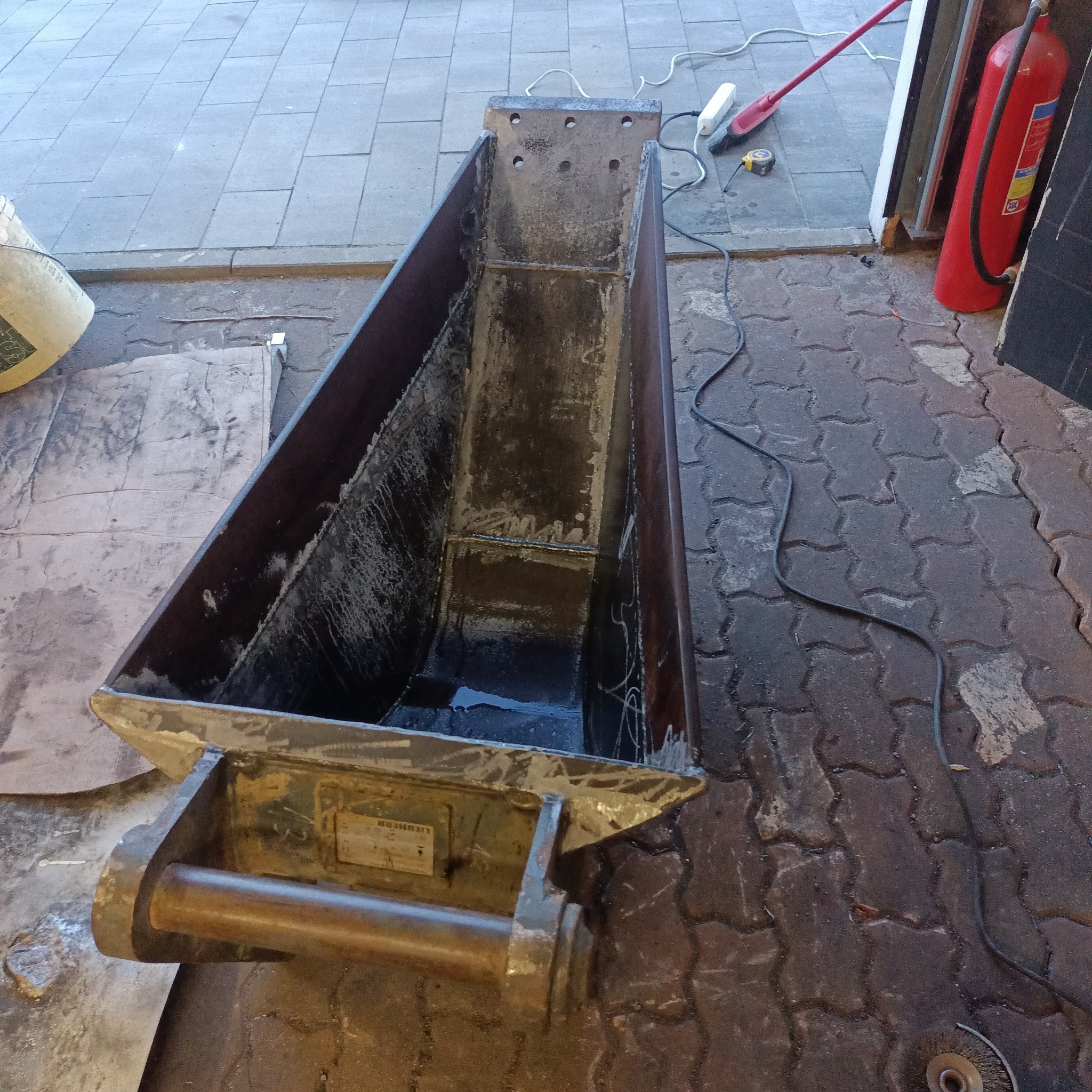 Metalowa skrzynia z widocznymi spawami, stojąca na brukowanej powierzchni, obok gaśnica i przewód zasilający