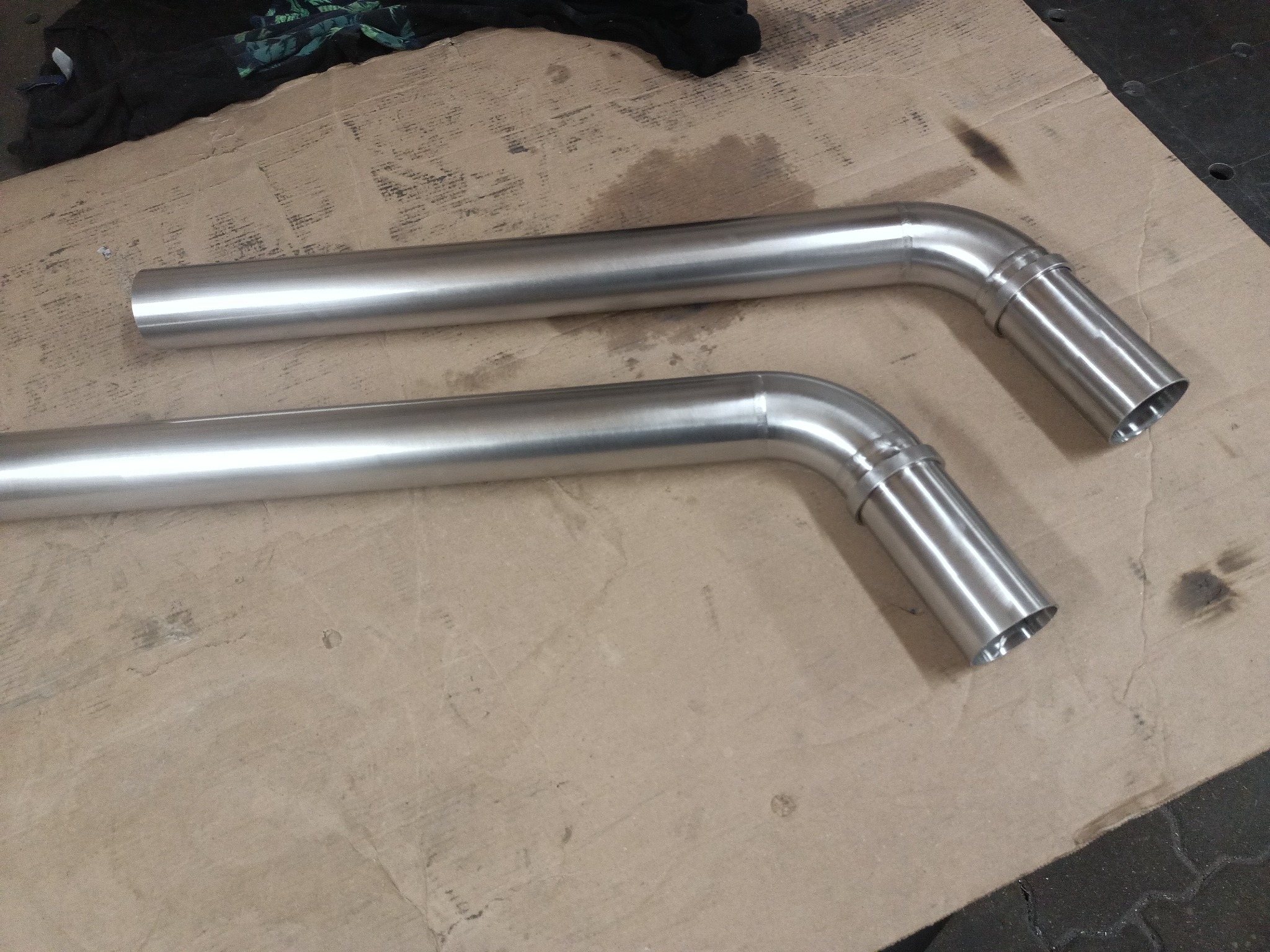 Dwie rury ze stali nierdzewnej, z kolankiem i widocznymi spawami, leżące na kartonowym podłożu.