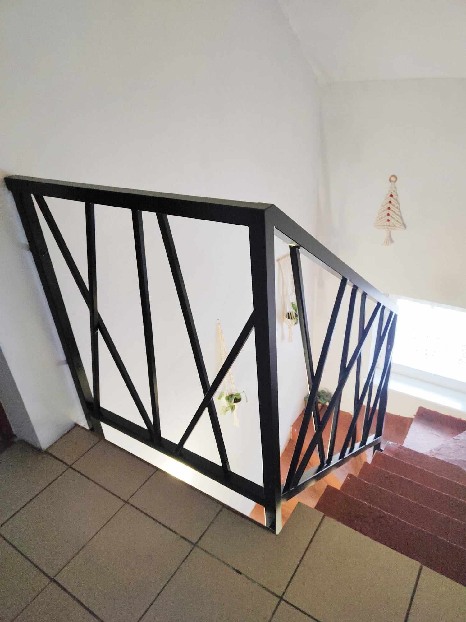 Nowoczesna, czarna balustrada schodowa z geometrycznym wzorem, widok z góry na klatkę schodową z terakotowymi schodami, dekoracje roślinne wiszące na balustradzie.