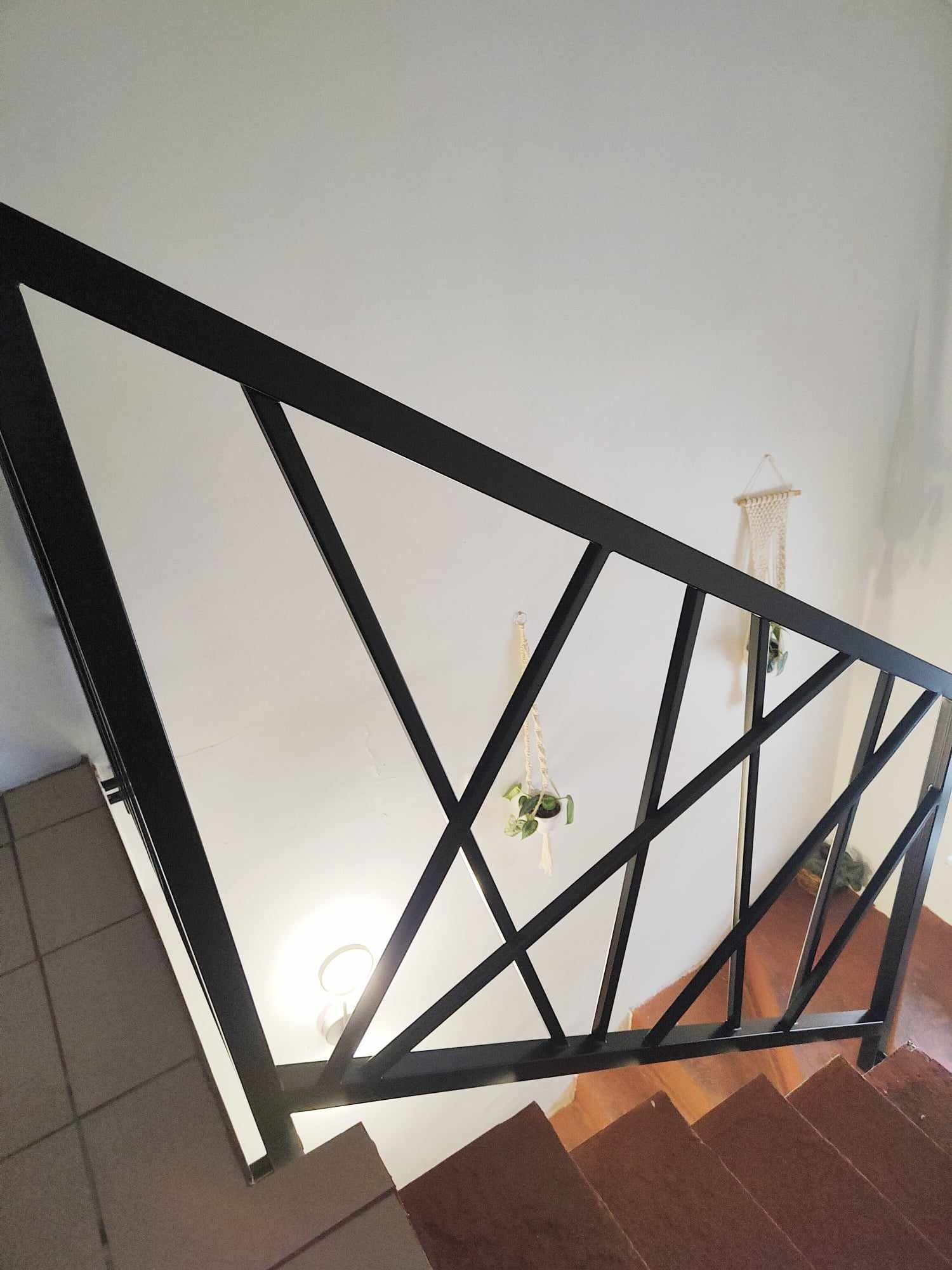 Nowoczesna, czarna balustrada schodowa z geometrycznym wzorem, widok z góry na drewniane stopnie i białe ściany z dekoracjami roślinnymi.