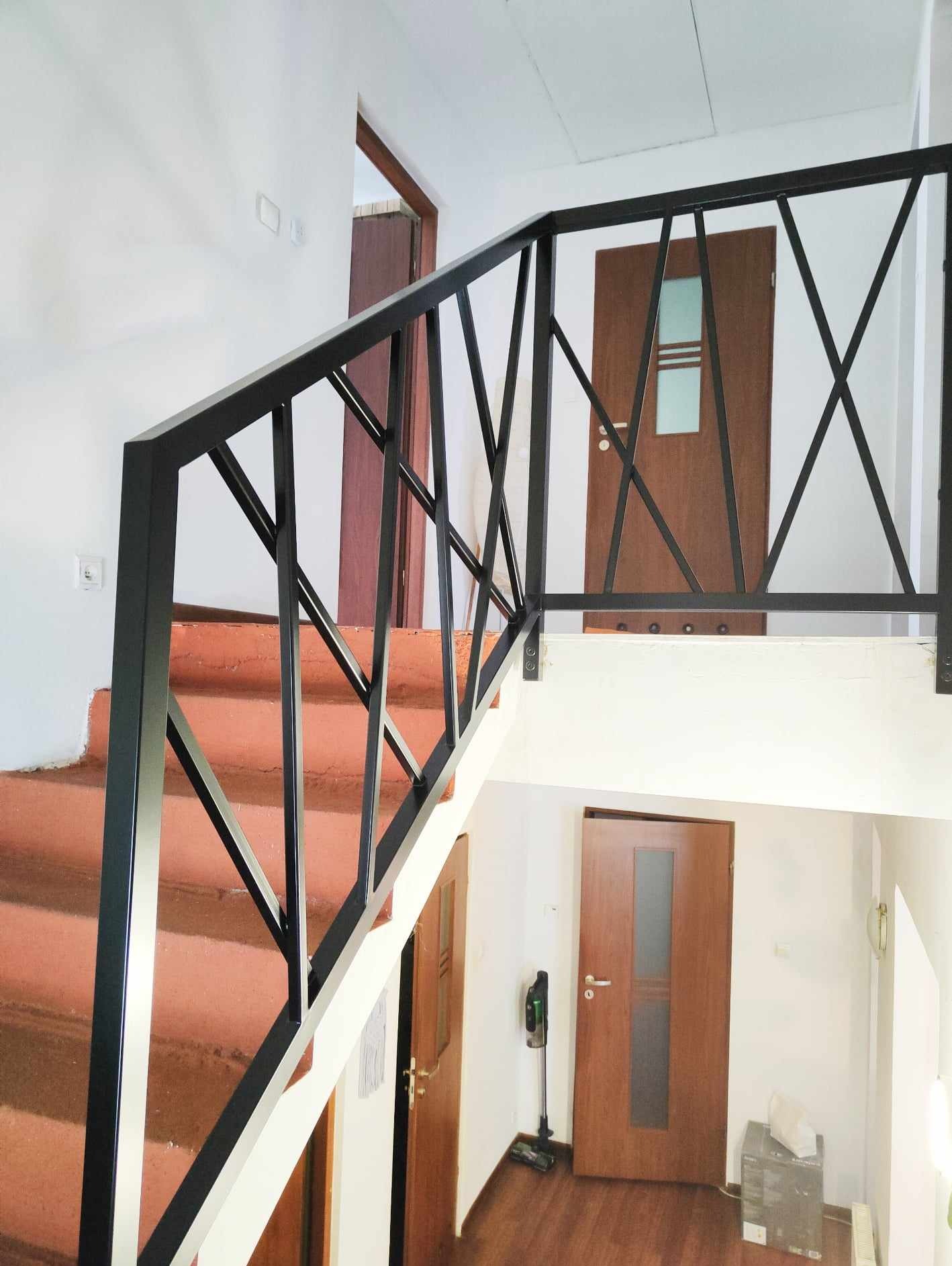 Klatka schodowa z nową, czarną, metalową balustradą o geometrycznym wzorze, widok z dołu na górę, z widocznymi drzwiami na różnych poziomach.