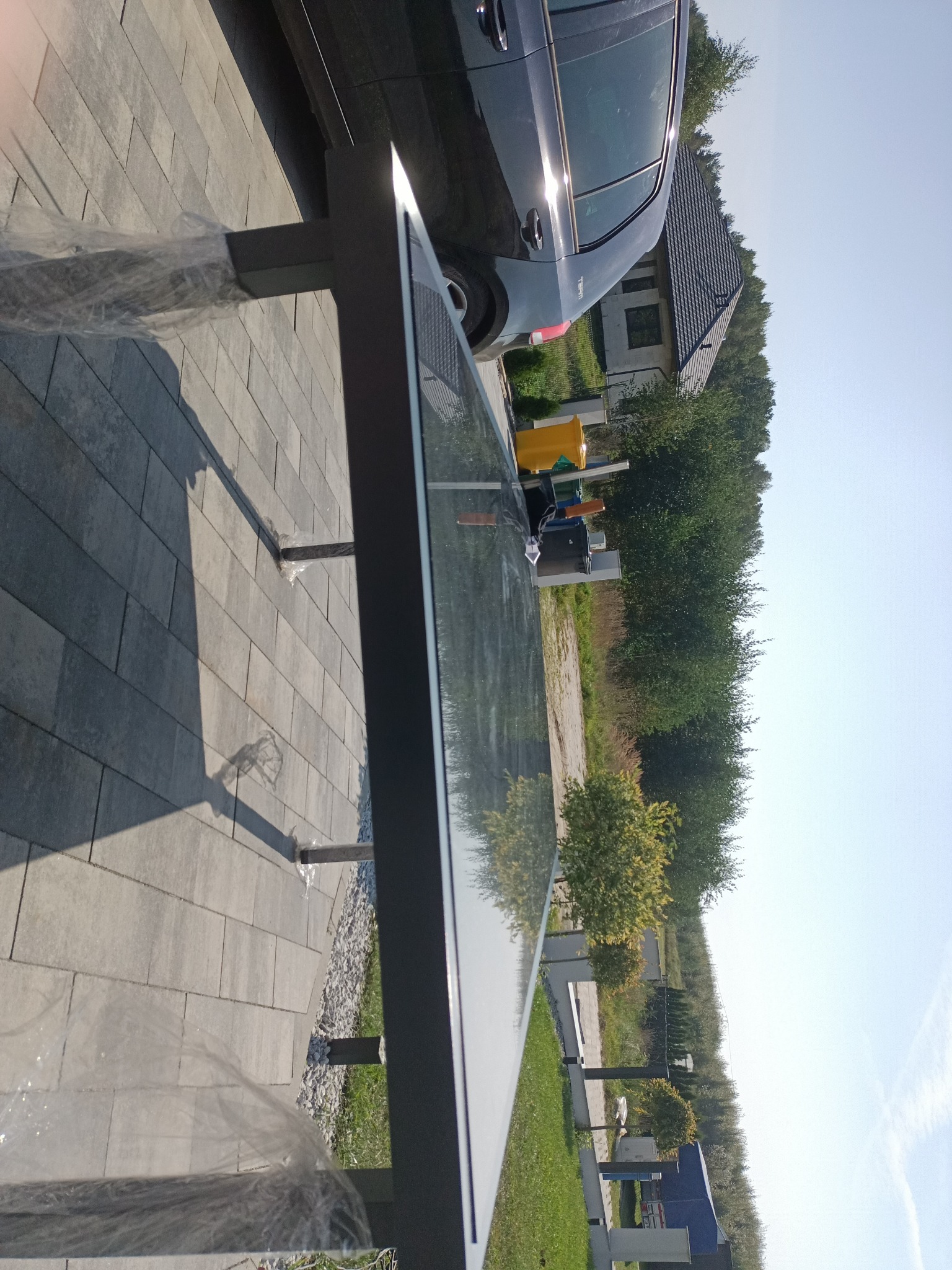 Szklany blat stołu z metalową ramą w folii ochronnej, na brukowanym tarasie, z widokiem na dom, samochód i zieleń.