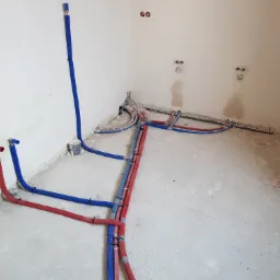 Rozprowadzenie instalacji wodnej i grzewczej w surowym stanie, widoczne rury w izolacji koloru niebieskiego i czerwonego, mocowane do ściany i podłogi za pomocą czarnych opasek.