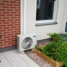Zewnętrzna jednostka klimatyzacji zamontowana na jasnych betonowych bloczkach na tarasie z szarych płyt, obok ceglanej ściany i drewnianej grządki z warzywami.