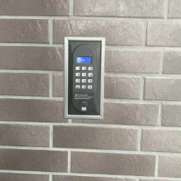 Panel domofonu cyfrowego ACO, zamontowany na ścianie z szarej cegły, wyświetlający napis 'Papr10' na niebieskim ekranie LCD.