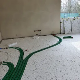 Wnętrze w trakcie instalacji ogrzewania podłogowego: zielone rury rozprowadzone na białych płytach izolacyjnych z wzorem lastryko, widoczne okno z zieloną siatką i płyty izolacyjne oparte o ścianę.