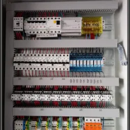 Otwarta szafa sterownicza z widocznymi zabezpieczeniami, przekaźnikami, listwami zaciskowymi i uporządkowanym okablowaniem w kolorach czerwonym, niebieskim i czarnym.