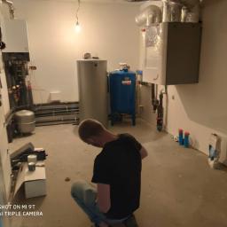 Blue Box Instalacje Sp. z o.o. - Instalator w czarnej koszulce kuca w kotłowni, gdzie widoczne są zbiorniki na wodę, rury, piec oraz narzędzia i materiały budowlane.
