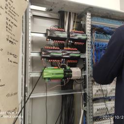 Blue Box Instalacje Sp. z o.o. - Panel sterowania inteligentnego domu w trakcie instalacji, widoczne moduły sterujące z podłączonymi przewodami, instalator w tle, po lewej stronie kartka z odręcznymi notatkami.