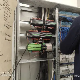 Panel sterowania inteligentnego domu w trakcie instalacji, widoczne moduły sterujące z podłączonymi przewodami, instalator w tle, po lewej stronie kartka z odręcznymi notatkami.