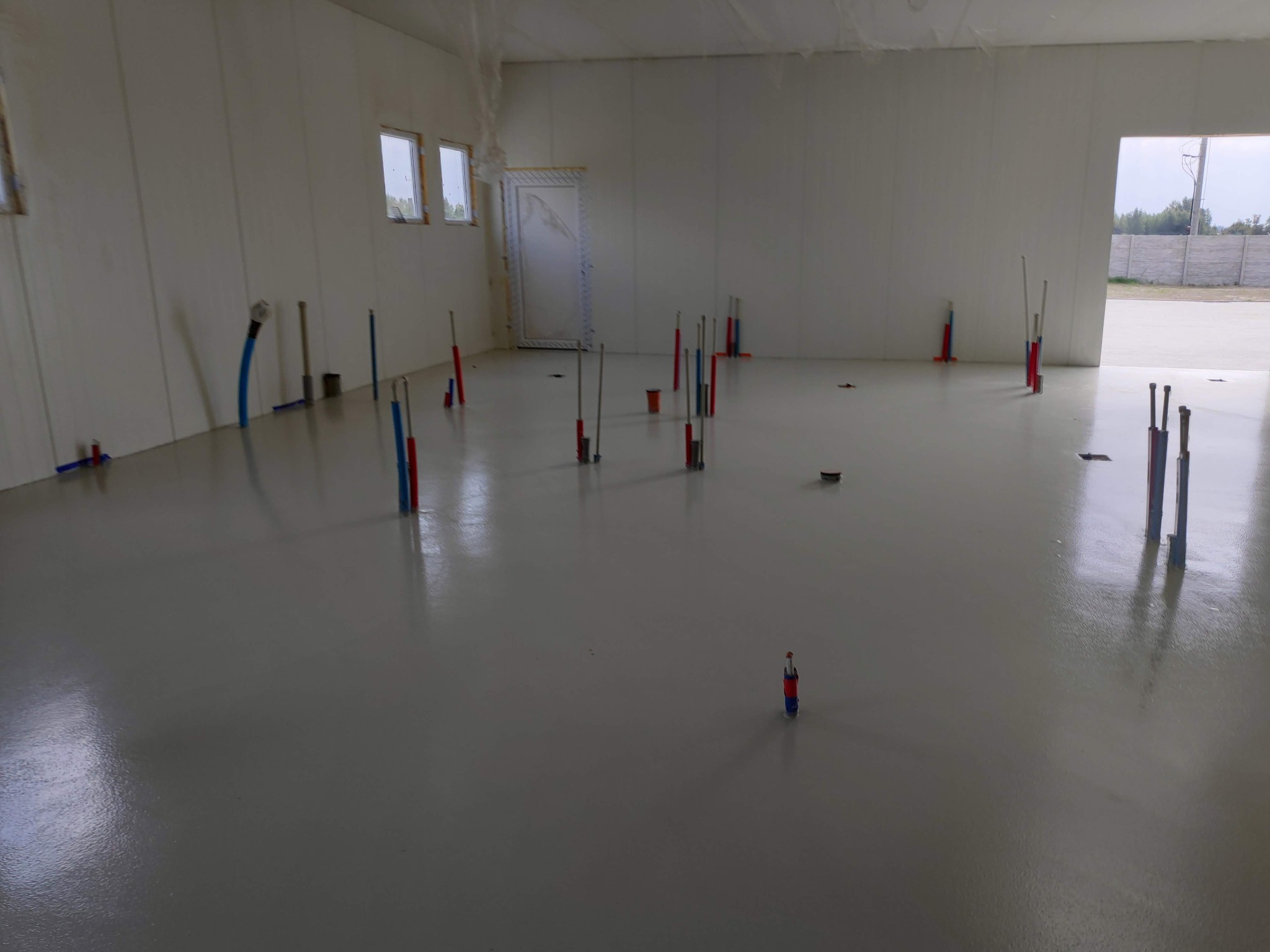 Wnętrze w trakcie instalacji ogrzewania podłogowego z widocznymi wystającymi rurami i przewodami, na wylewce samopoziomującej, w tle ściany z oknami.