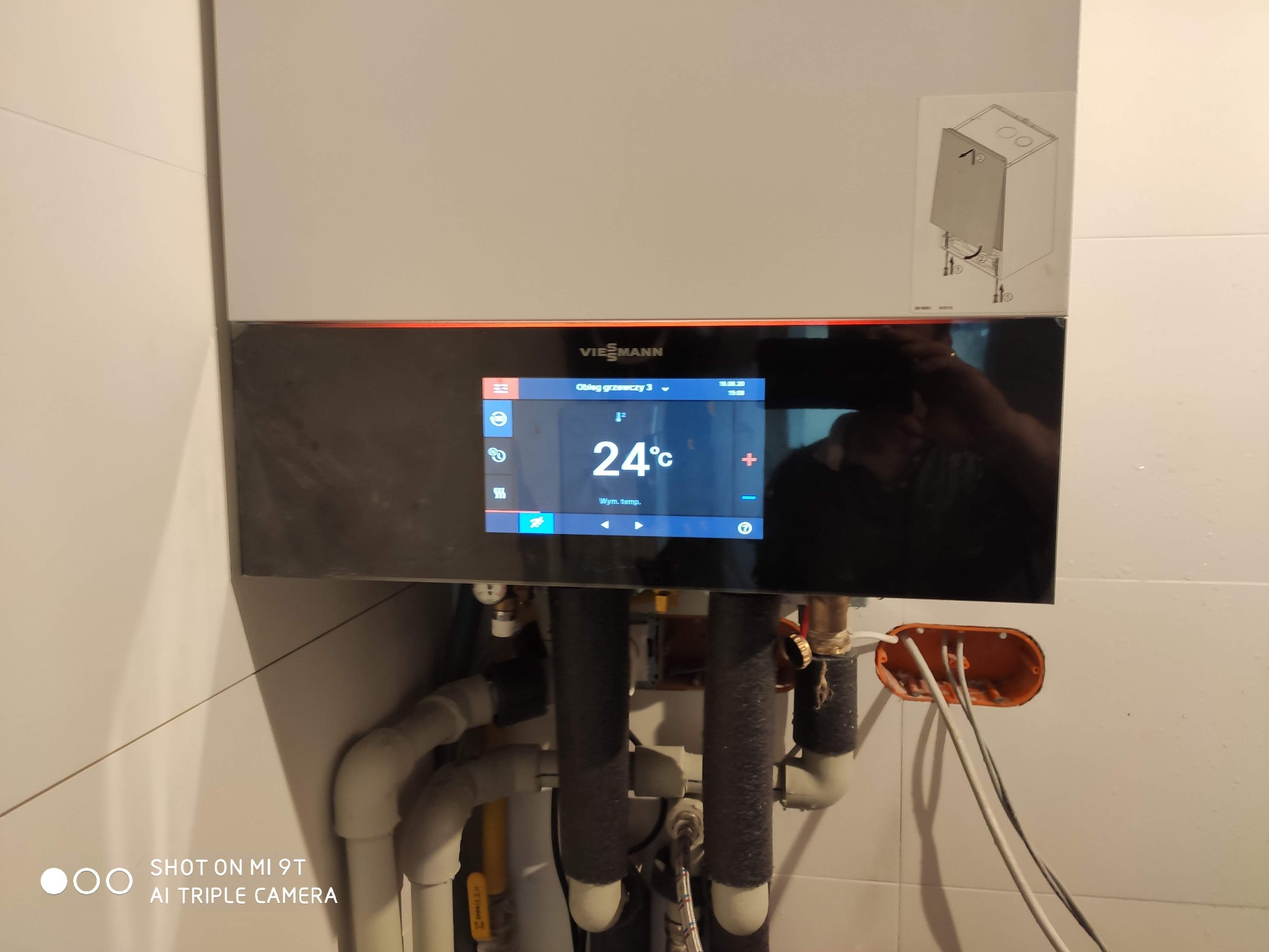 Nowoczesny piec gazowy Viessmann z cyfrowym wyświetlaczem pokazującym temperaturę 24°C, widoczne rury instalacji grzewczej i schemat montażowy na obudowie.