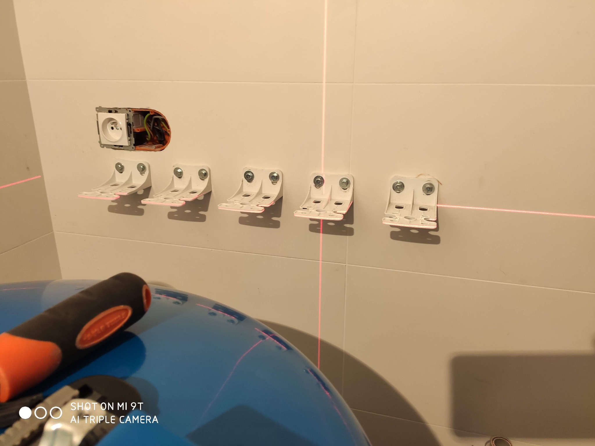 Montaż uchwytów na płytkach ściennych z użyciem poziomicy laserowej, widoczne gniazdko elektryczne i narzędzia na niebieskiej powierzchni.