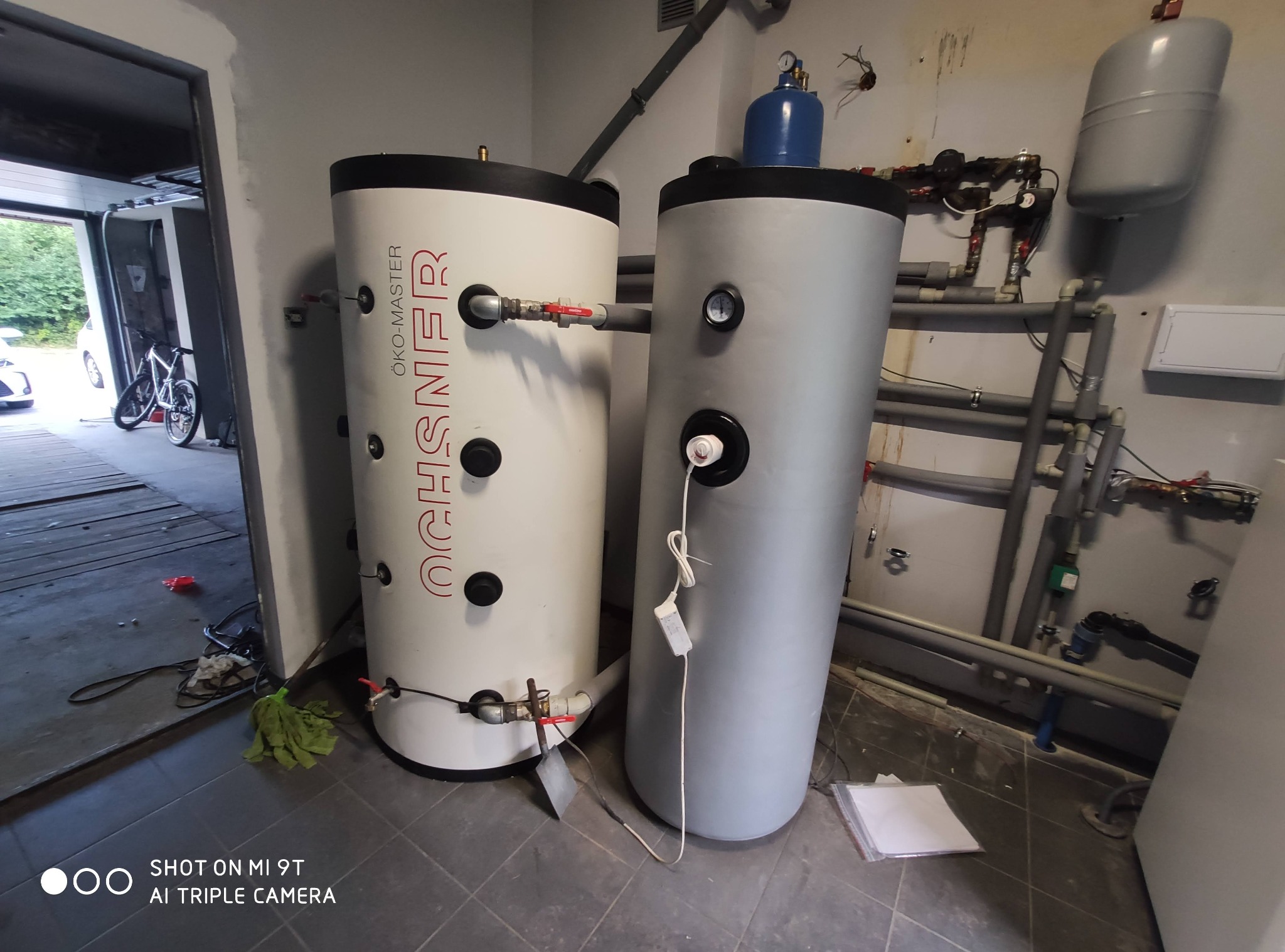 Dwa zbiorniki buforowe, biały z czerwonym napisem Öko-Master i szary, połączone instalacją hydrauliczną, w tle widoczne rury i osprzęt, na podłodze mop i dokumenty.