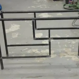 Metalowa konstrukcja stołu z rur o kwadratowym przekroju, spawana, surowa, bez blatu, widoczne spawy, na betonowej podłodze, w tle narzędzia.