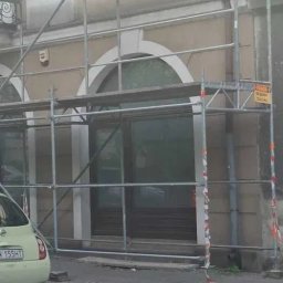 OMBUD Sławomir Mendera - Elewacja budynku w trakcie prac remontowych, widoczne rusztowania i fragment zaparkowanego samochodu. Detal architektoniczny: łukowe okna.