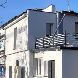 Dwukondygnacyjny budynek po remoncie elewacji z balkonem, widoczne świeże tynki i elementy wykończeniowe, rura spustowa na pierwszym planie, na drugim planie rusztowanie.