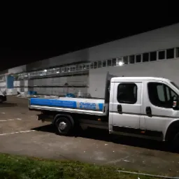 Dwa białe samochody dostawcze firmy budowlanej OMBUD na placu budowy w nocy; w tle budynek w trakcie prac elewacyjnych, widoczne rusztowania i materiały budowlane.