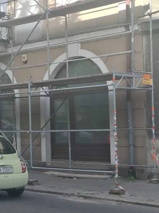 Elewacja budynku w trakcie prac remontowych, widoczne rusztowania i fragment zaparkowanego samochodu. Detal architektoniczny: łukowe okna.