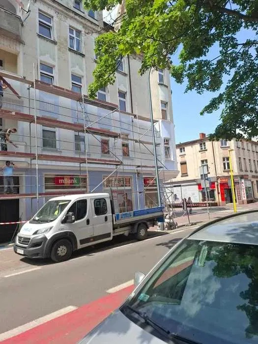 Prace na wysokościach: rusztowania przy elewacji kamienicy, biały samochód dostawczy na ulicy, fragment zaparkowanego auta w kadrze. Dzień słoneczny.