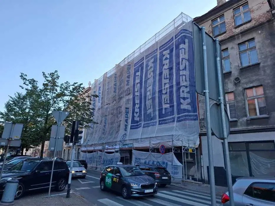 Budynek w rusztowaniach, osłonięty siatką z logo producentów materiałów budowlanych, widok z ulicy z przejeżdżającymi samochodami i sygnalizacją świetlną.
