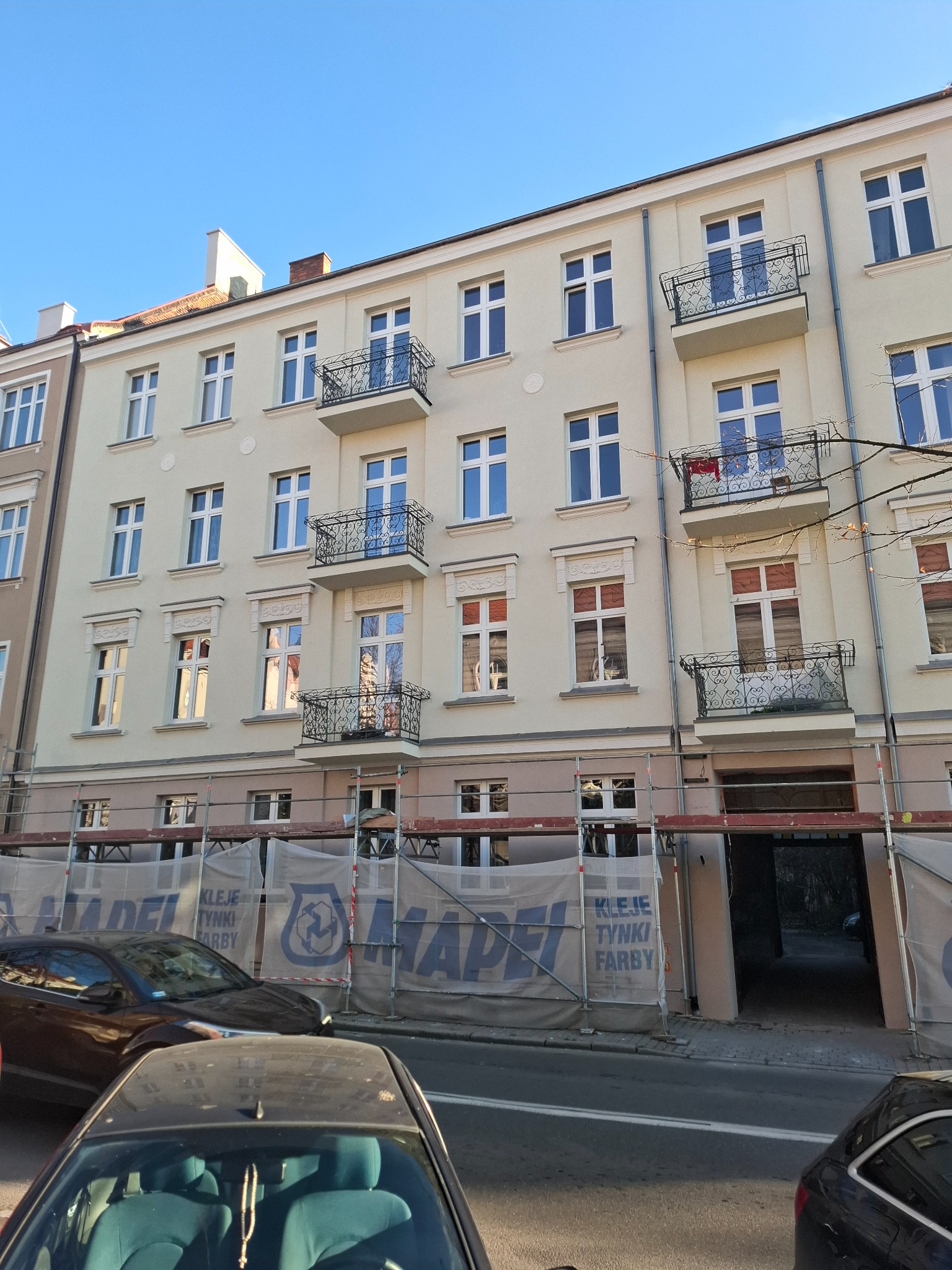 Elewacja kamienicy w trakcie prac. Widoczne rusztowania z siatką ochronną z logo producenta materiałów budowlanych. Balkony z ozdobnymi balustradami.