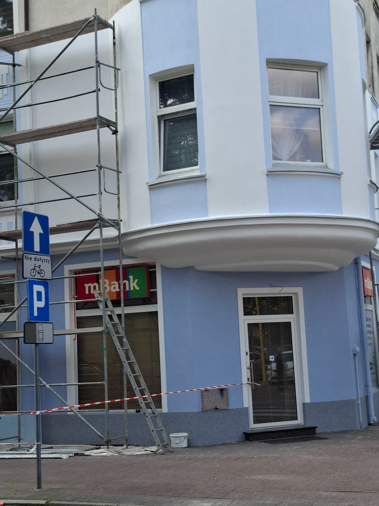 Narożny budynek po renowacji elewacji w biało-niebieskie pasy. Widoczne rusztowania, znak drogowy i logo banku. Ujęcie z dołu, podkreślające architekturę.
