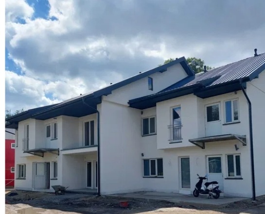 Nowoczesny budynek wielorodzinny z białą elewacją i ciemnym dachem. Widoczny skuter i taczka, sugerujące prace wykończeniowe. Ujęcie w słoneczny dzień.