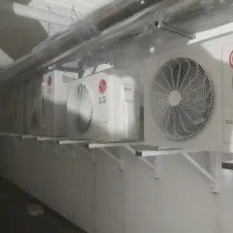 Rząd białych jednostek zewnętrznych klimatyzacji LG Dual Inverter zamontowanych na wspornikach na ścianie w garażu, widoczne rury wentylacyjne pod sufitem.