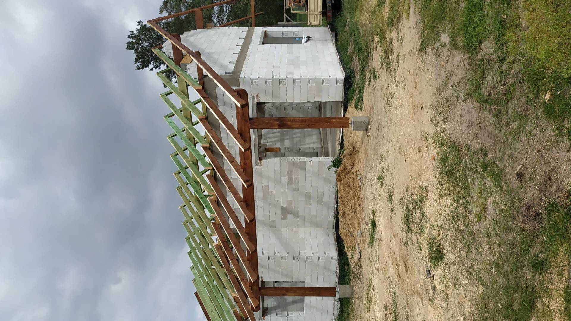 Budowa domu z bloczków: widoczna konstrukcja drewniana dachu z zielonymi elementami i fragment budynku w surowym stanie pod pochmurnym niebem.