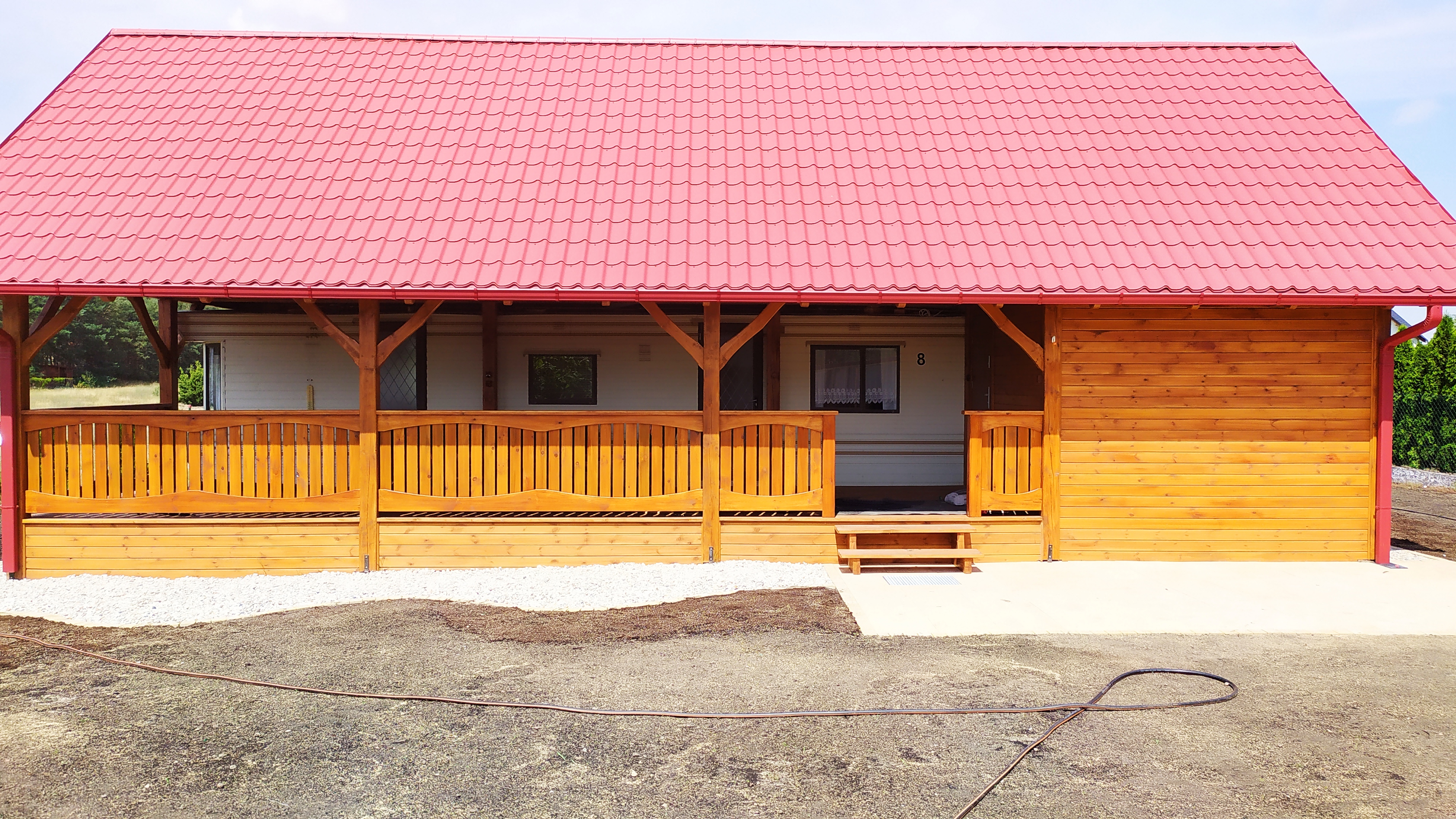 Nowo wybudowany domek letniskowy z czerwonym dachem z blachodachówki, drewnianą elewacją i werandą z balustradą, widok z przodu na tle błękitnego nieba.