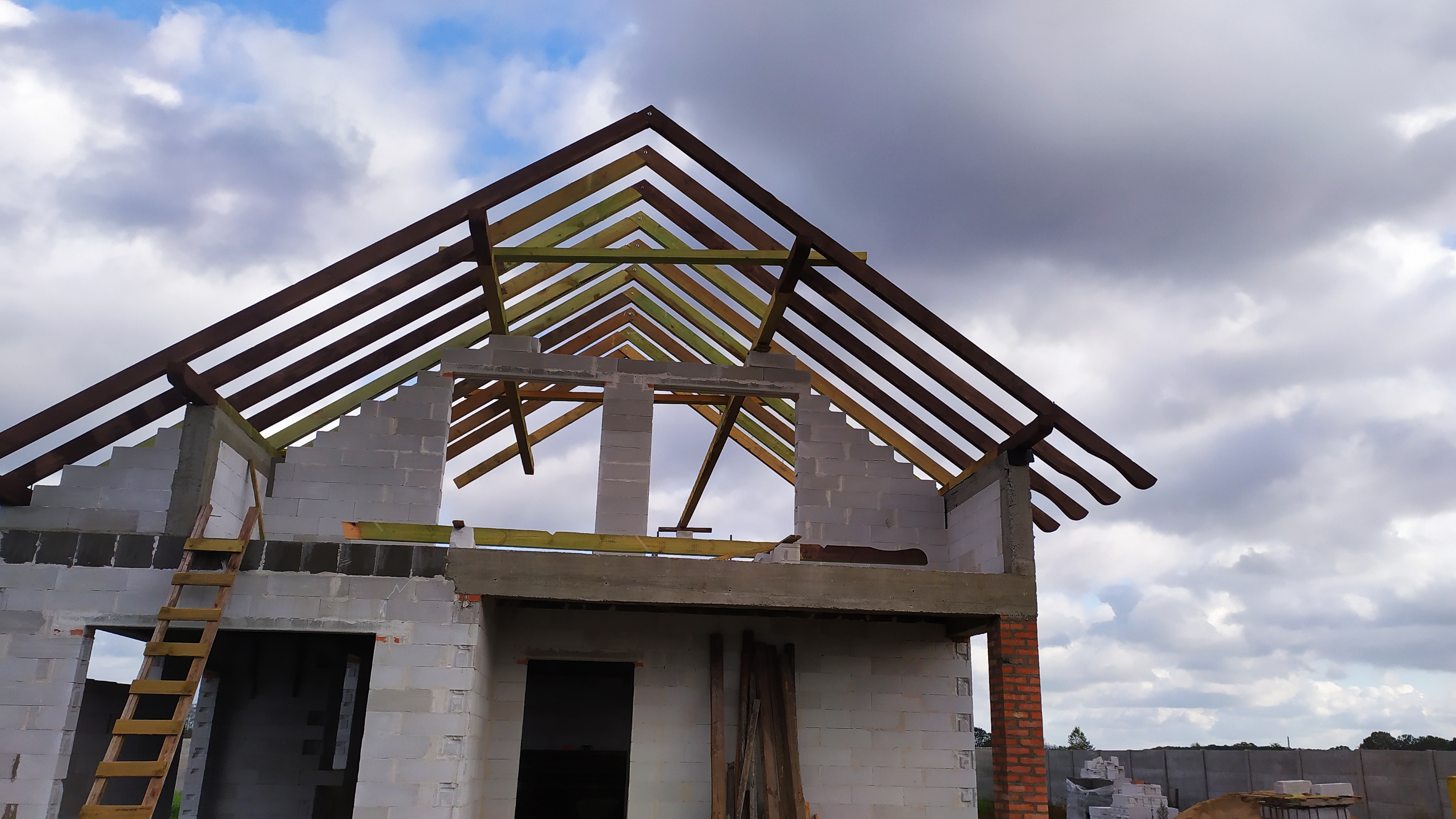 Wznoszenie więźby dachowej na surowym budynku z betonu komórkowego, widoczne drewniane elementy konstrukcyjne dachu i drabina oparta o mur.