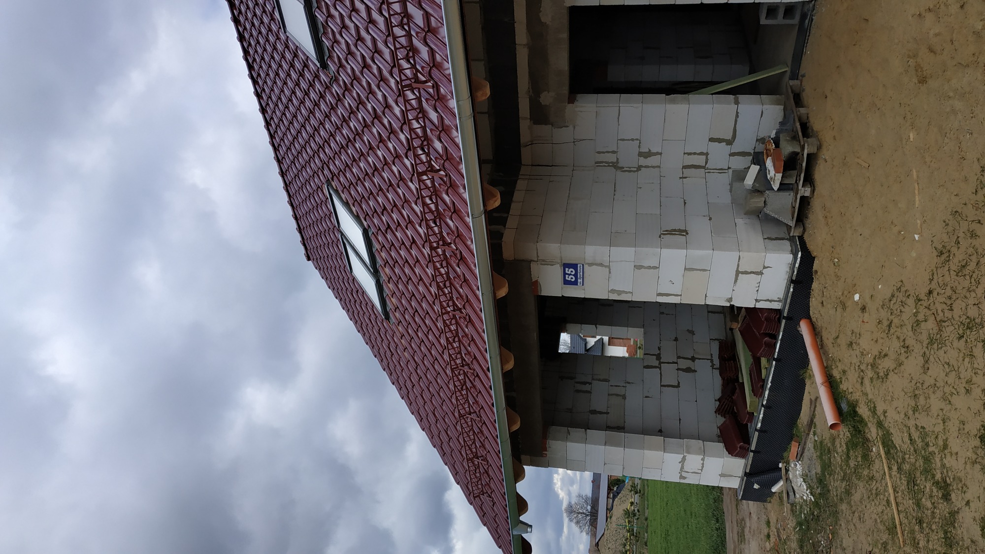 Czerwony dach z dachówki ceramicznej na surowym budynku z białych pustaków, widok z dołu na tle pochmurnego nieba.